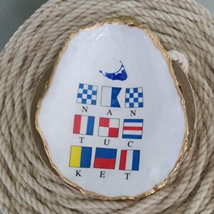 May include: White oyster shell dish with a gold rim, featuring nautical signal flags and the word "NAN" and "KET". The dish has a blue and white design of a cape. A decorative coastal accent.