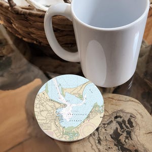 May include: A white ceramic mug rests on a wooden surface next to a round coaster. The coaster features a nautical chart of Nantucket Harbor, with blue and beige tones. A woven basket is partially visible in the background.