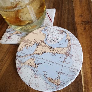 May include: Two ceramic coasters with a nautical map design. One coaster is round, and the other is square. A glass with ice and a light-colored liquid sits on top of the coasters. The coasters feature shades of blue, beige, and white.