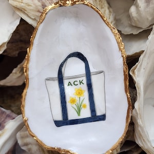 May include: A white oyster shell dish with a gold-painted rim, featuring a hand-painted illustration of a white tote bag with dark blue handles and trim. The bag displays the text ACK in green letters above a cluster of three yellow daffodils.