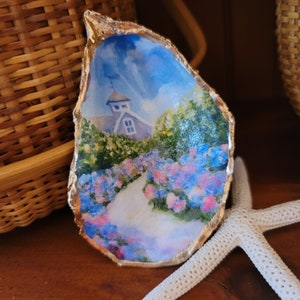 May include: A small, gold-leafed oyster shell with a painted scene of a white house with a blue roof and a path lined with pink and blue flowers. The background is a blue sky with white clouds.
