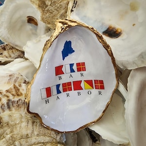May include: An oyster shell with a gold-painted rim and a white interior. The shell is decorated with a blue Maine outline and the words "BAR HARBOR" spelled out with nautical signal flags.