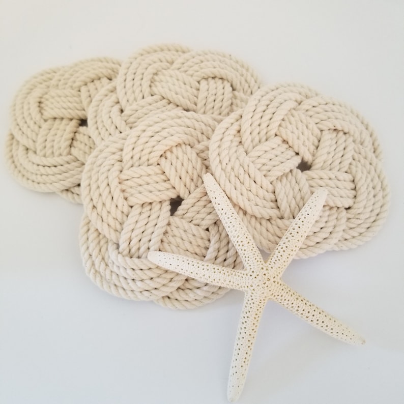K&ouml;nnte beinhalten: Vier cremefarbene Seiluntersetzer mit einem geflochtenen Knotendesign und einem wei&szlig;en Seestern.