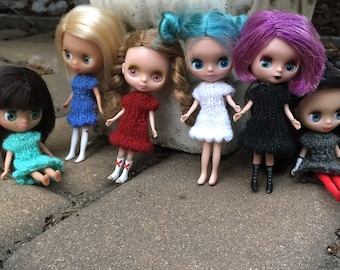 May include: Seven dolls wearing knitted dresses in various colors, including blue, red, white, black, and gray. The dolls are posed on a gray tiled surface.