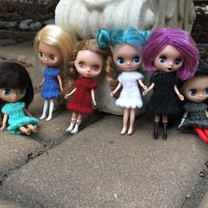 May include: Seven dolls wearing knitted dresses in various colors, including blue, red, white, black, and gray. The dolls are posed on a gray tiled surface.
