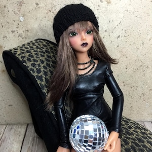 May include: A doll wearing a black knit beanie, a black leather dress, and a silver disco ball. The doll has long brown hair and green eyes.