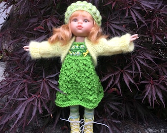 May include: A doll wearing a green knitted dress with a yellow cardigan and a green and white hat. The doll has blonde hair and blue eyes.