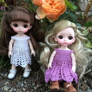 May include: Two dolls wearing crocheted dresses. The doll on the left has brown hair and is wearing a white dress. The doll on the right has blonde hair and is wearing a purple dress.