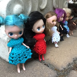 May include: Six dolls with long hair and crocheted dresses. The dolls are standing in a row on a gray surface. The dolls are wearing dresses in various colors, including red, white, blue, and turquoise.