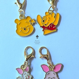 Könnte beinhalten: Vier goldfarbene Charm-Clips mit Zeichentrickfiguren. Zwei Anhänger zeigen einen gelben Bären mit rotem Hemd, und die anderen beiden Anhänger zeigen ein rosa Schwein. Die Anhänger sind an goldfarbenen Verschlüssen befestigt.