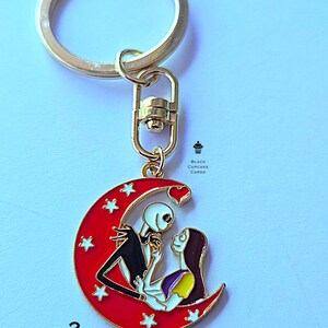 May include: A gold-colored keychain with a red crescent moon pendant featuring Jack and Sally from The Nightmare Before Christmas. The pendant has white stars and the number 2.