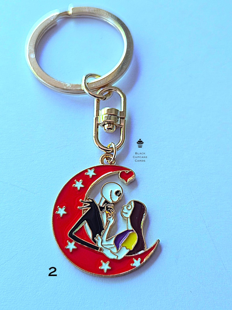 May include: A gold-colored keychain with a red crescent moon pendant featuring Jack and Sally from The Nightmare Before Christmas. The pendant has white stars and the number 2.