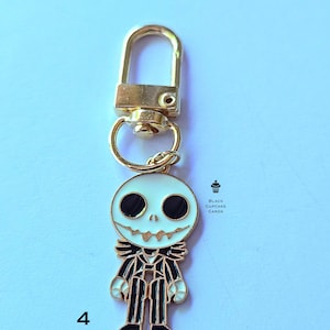 May include: A gold-colored metal keychain featuring a white and black Jack Skellington character design. The character has large black eyes and a stitched grin. The keychain includes a lobster clasp and a split ring. The text "Black Cupcake Cards" is visible.