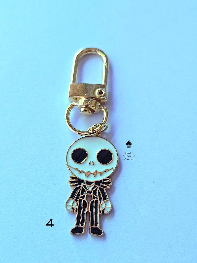May include: A gold-colored metal keychain featuring a white and black Jack Skellington character design. The character has large black eyes and a stitched grin. The keychain includes a lobster clasp and a split ring. The text "Black Cupcake Cards" is visible.