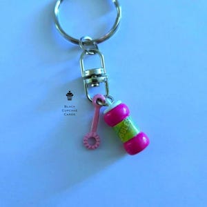 May include: A keychain with a silver ring and clasp, featuring a miniature pink bubble wand and a pink and green bubble solution bottle. The text "Bubbles" is visible on the bottle. The logo "Black Cupcake Cards" is also present.