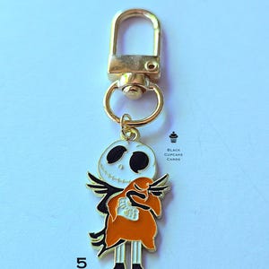 May include: A gold-toned keychain featuring a Jack Skellington charm. The charm is white with black eyes and a stitched smile, wearing a black suit and orange coat. The keychain has a lobster clasp and a small ring.
