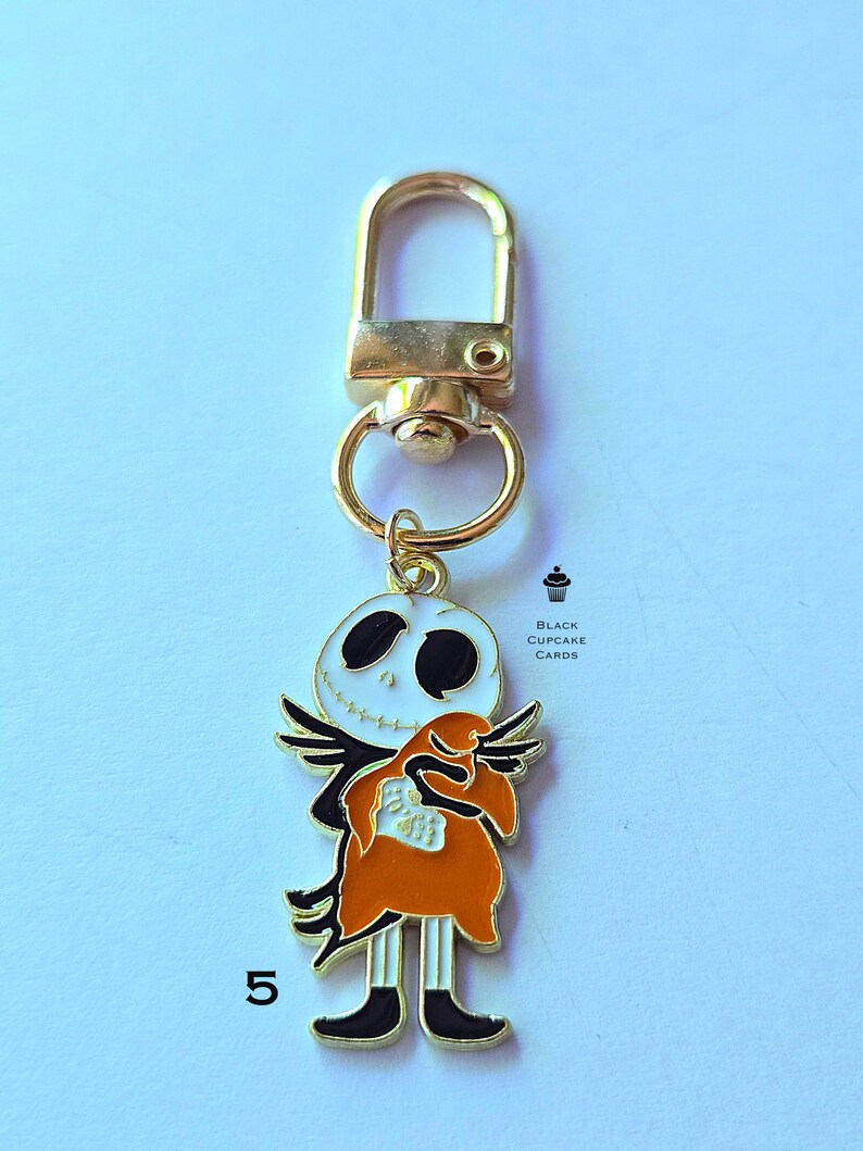 May include: A gold-toned keychain featuring a Jack Skellington charm. The charm is white with black eyes and a stitched smile, wearing a black suit and orange coat. The keychain has a lobster clasp and a small ring.
