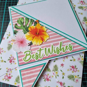 May include: A handmade greeting card with a pink and white striped background, a green leaf, and a pink and yellow hibiscus flower. The card is folded into a triangle shape and has the words "Best Wishes" written on it.
