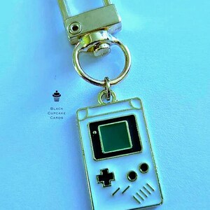 May include: A white and gold keychain featuring a miniature retro handheld game console design. The keychain has a gold clasp and ring, with a white enamel game console pendant. The game console has a black screen and buttons, with the text "Black Cupcake Cards".