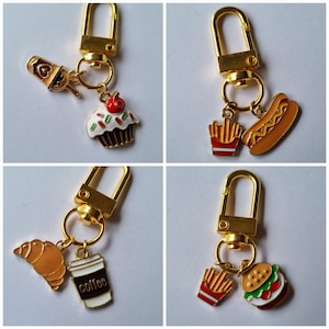May include: Four gold keychains with different food-themed charms. The charms include a cupcake, a hot dog, a coffee cup, and a croissant. Each charm is attached to a gold ring and a gold chain.