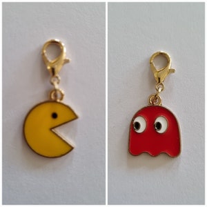 Op de afbeelding: Twee gouden bedels, een in de vorm van Pac-Man met een geel gezicht en zwarte ogen, en de andere in de vorm van een spook met een rood gezicht en witte ogen.