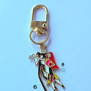 May include: Gold-colored keychain featuring a charm with Jack Skellington and Sally from The Nightmare Before Christmas. The charm is colorful with black, white, red, pink, and yellow details. The number 6 is printed on the bottom left.