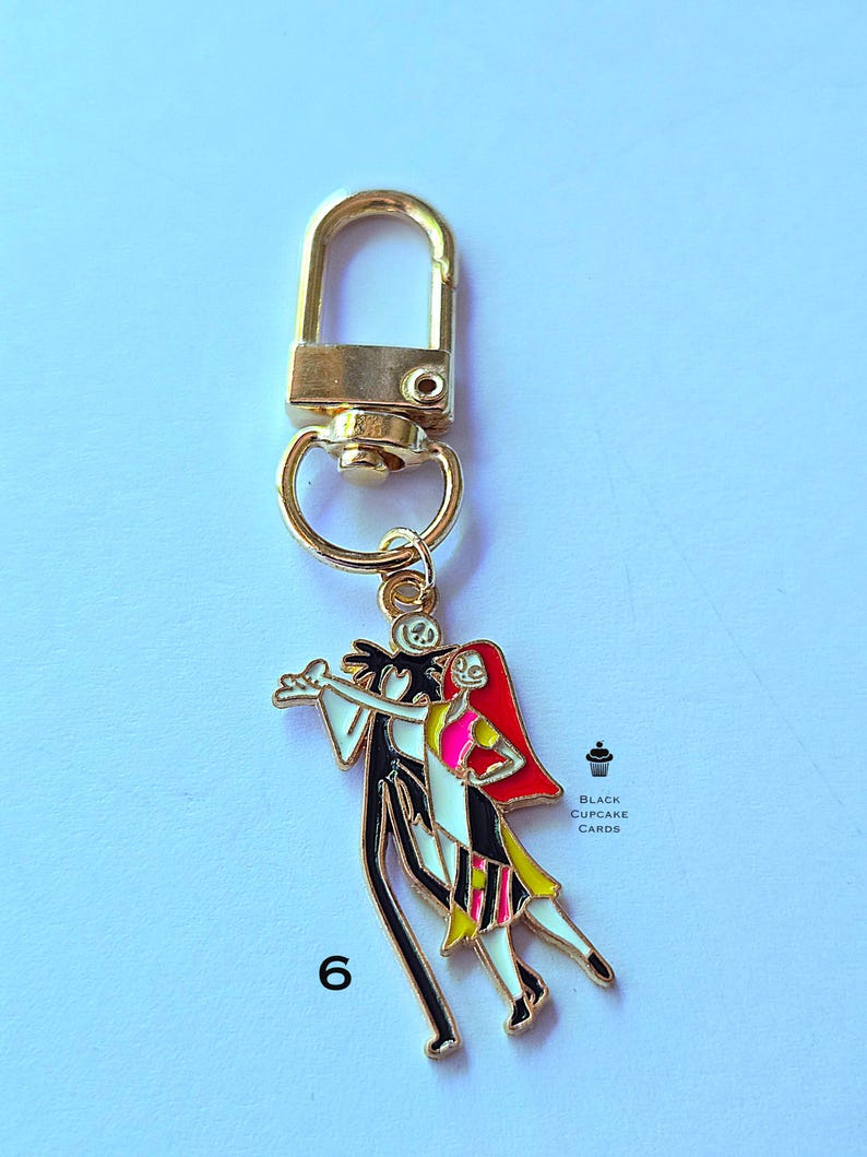 May include: Gold-colored keychain featuring a charm with Jack Skellington and Sally from The Nightmare Before Christmas. The charm is colorful with black, white, red, pink, and yellow details. The number 6 is printed on the bottom left.