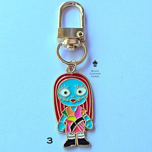 May include: A gold-colored metal keychain featuring a cartoon character with blue skin, red hair, and a patchwork dress. The character has stitched-up facial features. The number "3" is visible on the bottom left. The keychain has a lobster clasp.