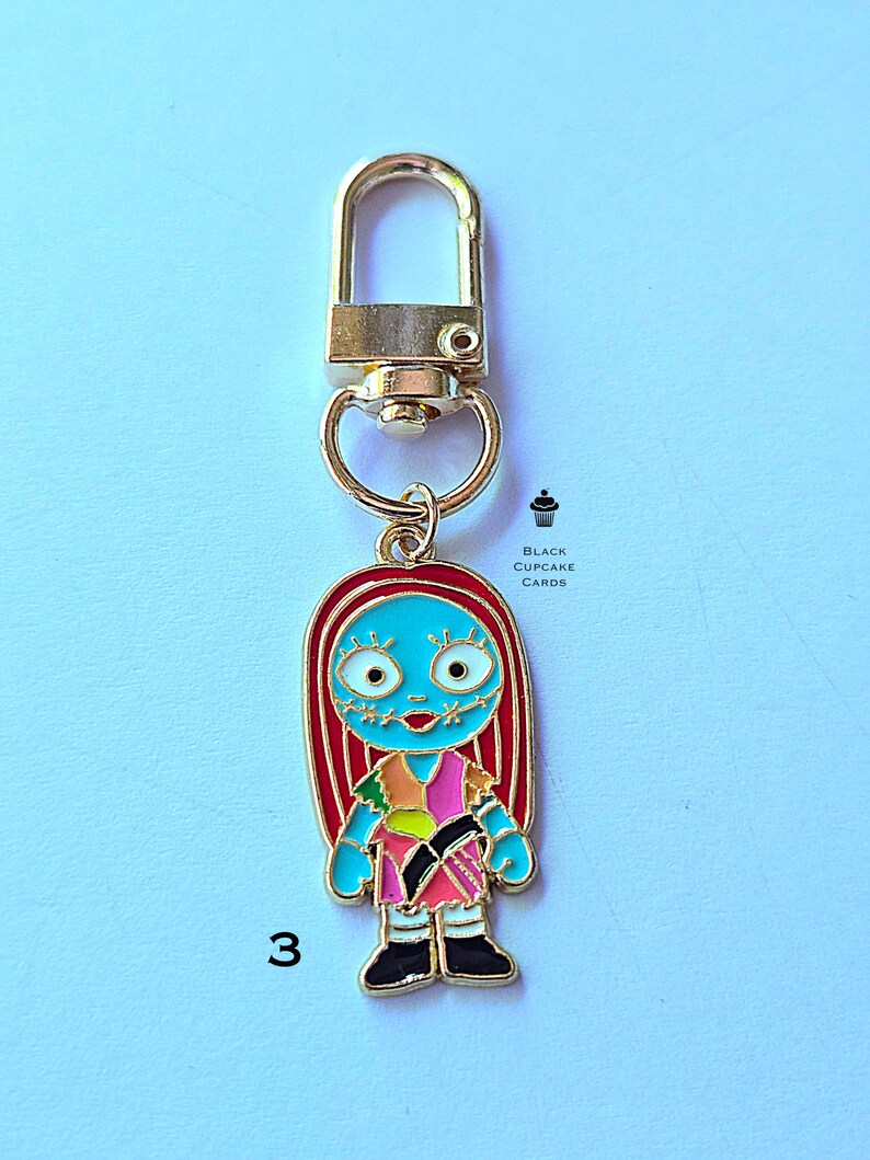 May include: A gold-colored metal keychain featuring a cartoon character with blue skin, red hair, and a patchwork dress. The character has stitched-up facial features. The number "3" is visible on the bottom left. The keychain has a lobster clasp.