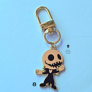 May include: Gold-toned metal keychain featuring a cartoon skeleton character with a round head, black eyes, and a jagged smile. The skeleton is wearing a black outfit. The keychain has a gold clasp and ring. The number 8 is printed on the bottom left.