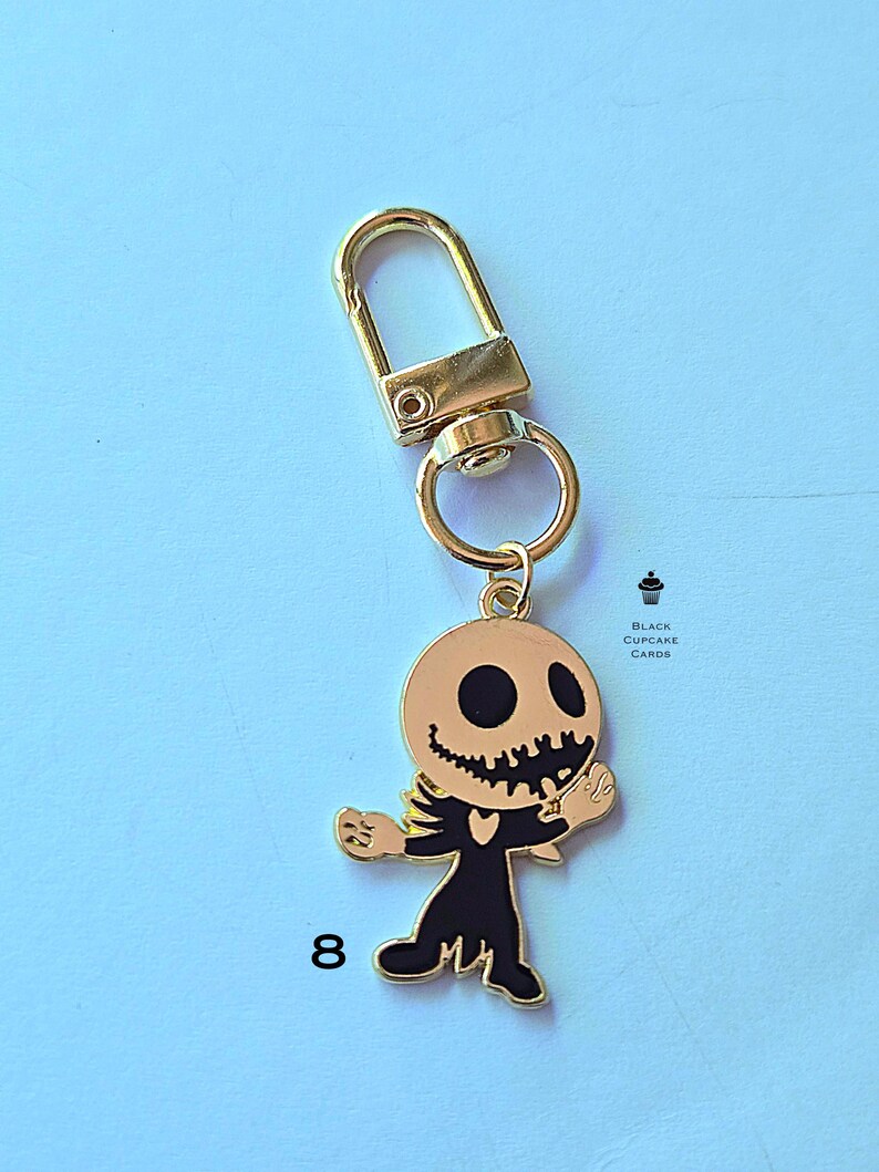 May include: Gold-toned metal keychain featuring a cartoon skeleton character with a round head, black eyes, and a jagged smile. The skeleton is wearing a black outfit. The keychain has a gold clasp and ring. The number 8 is printed on the bottom left.