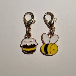 Puede incluir: Dos abalorios de metal dorado con diseños de abejas. Un abalorio presenta un tarro de miel con una tapa blanca y rayas amarillas y negras. El otro abalorio presenta una abeja de dibujos animados con alas blancas, un cuerpo amarillo y rayas negras.