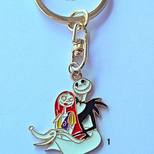 May include: A gold-colored metal keychain featuring a charm with Jack Skellington and Sally from The Nightmare Before Christmas. The charm is white, black, red, and yellow. The text "BLACK CUPCAKE CARDS" is printed on the keychain.