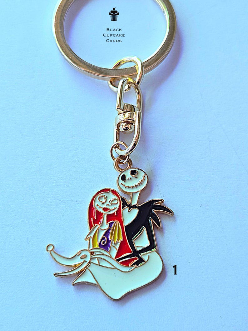 May include: A gold-colored metal keychain featuring a charm with Jack Skellington and Sally from The Nightmare Before Christmas. The charm is white, black, red, and yellow. The text "BLACK CUPCAKE CARDS" is printed on the keychain.