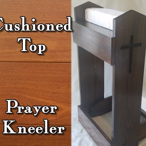 May include: A dark stained wooden prayer kneeler with a cushioned top and knee rest. A black cross is carved into the back of the kneeler. The text "Cushioned Top" and "Prayer Kneeler" are visible on the left side of the image.