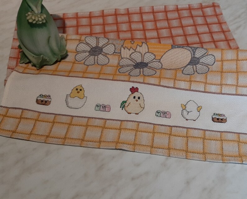 K&ouml;nnte beinhalten: Ein wei&szlig;es K&uuml;chentuch mit einem gelben und orangefarbenen Karomuster. Das Tuch zeigt gestickte Bilder von K&uuml;ken, Eiern und einem Korb mit Eiern.