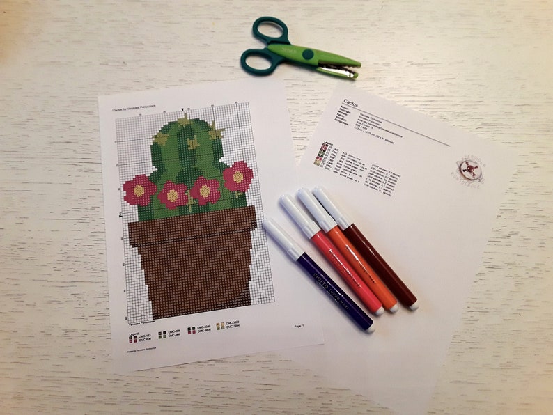 Op de afbeelding: Een kruissteekpatroon voor een cactus in een pot met roze bloemen. Het patroon is afgedrukt op wit papier en bevat een kleurenkaart en instructies. Er liggen ook vijf gekleurde stiften op de tafel.