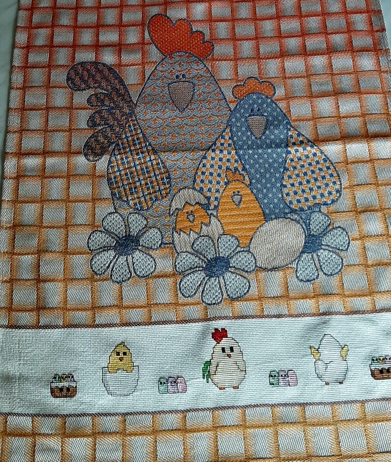 K&ouml;nnte beinhalten: Ein K&uuml;chentuch mit einem bunten gestickten Design mit zwei H&uuml;hnern und K&uuml;ken. Die H&uuml;hner sind blau und grau mit orangefarbenen K&auml;mmen und die K&uuml;ken sind gelb. Der Hintergrund ist ein kariertes Muster aus Orange und Wei&szlig;.