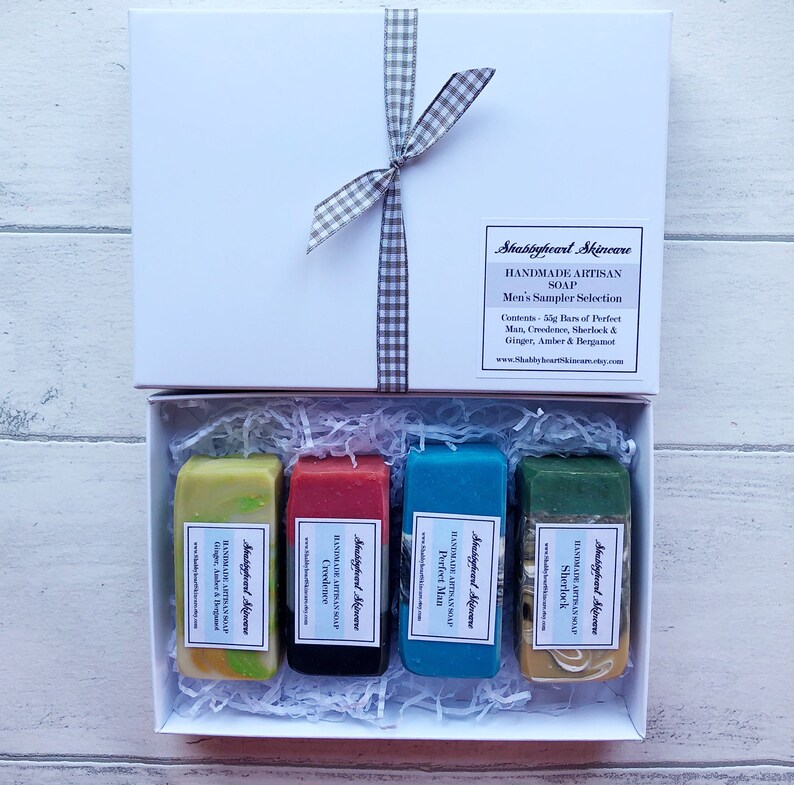 May include: A white gift box with a gray and white checkered ribbon. The box contains four bars of handmade soap. The soap bars are labeled with the names "Perfect Man", "Creedence", "Sherlock", and "Ginger, Amber & Bergamot".