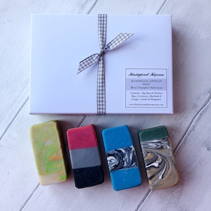 May include: A white gift box with a gray and white checkered ribbon tied around it. The box contains four bars of handmade artisan soap. The soap bars are in various colors and patterns, including green, red, blue, and black.