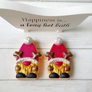 May include: Two gnome-shaped bath bombs with red hats, gold bodies, and brown antlers. The bath bombs have black shoes and white trim. Above them is a white bathtub-shaped sign with the text "Happiness is... a long hot bath."