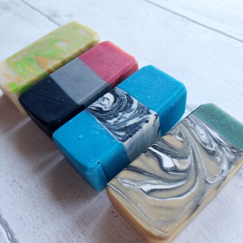 May include: Four bars of handmade soap with different colors and patterns. The soaps are arranged in a row from left to right. The first soap is green and yellow with a swirl pattern. The second soap is black and gray with a swirl pattern. The third soap is blue and white with a swirl pattern. The fourth soap is beige and white with a swirl pattern.