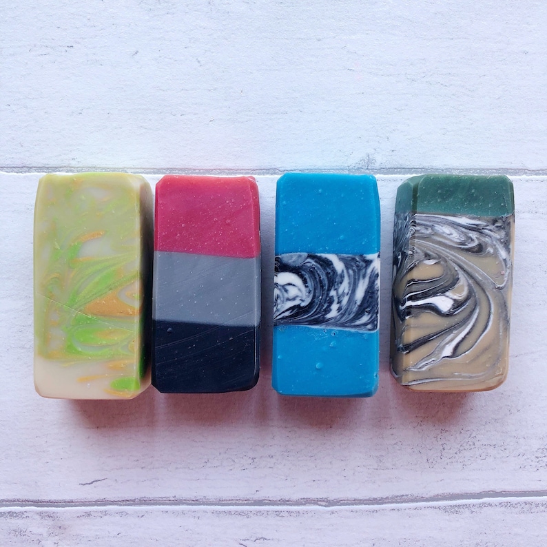May include: Four bars of handmade soap with different color combinations and swirl patterns. The soaps are arranged in a row from left to right: green and yellow, red and gray, blue and black, and beige and brown.
