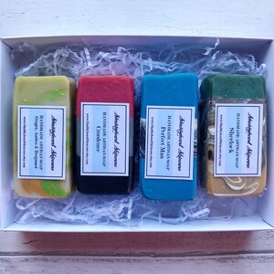 May include: Four bars of handmade artisan soap in a white box with shredded paper. The soaps are labeled with the names "Sherlock", "Perfect Man", "Creedence", and "Ginger, Amber & Bergamot".