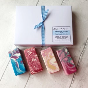 May include: A white gift box tied with a blue and white gingham ribbon, containing four bars of handmade artisan soap. The soap bars are in a variety of colours and swirl patterns, including blue, pink, and red.