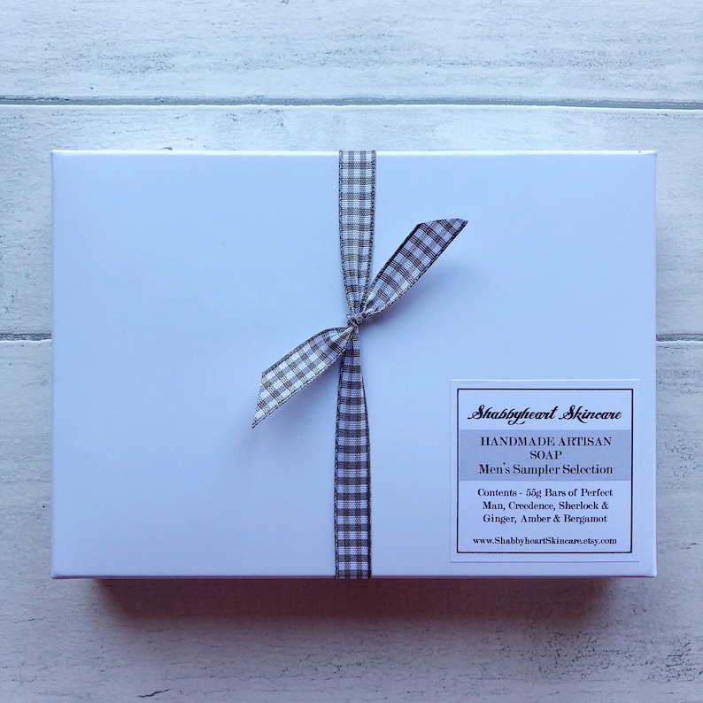 May include: A white gift box with a black and white checkered ribbon tied around it. The box has a label that reads "Shabbyheart Skincare Handmade Artisan Soap Men's Sampler Selection Contents - 50g Bars of Perfect Man, Creedence, Sherlock & Ginger, Amber & Bergamot www.ShabbyheartSkincare.etsy.com"