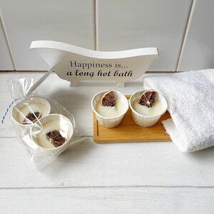May include: A spa-themed image featuring white bath bombs with dried orange slices, displayed in paper cups. A white bathtub-shaped sign reads "Happiness is... a long hot bath." A white towel and a wooden tray are also present.