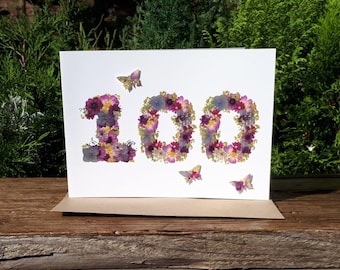 May include: A white card with the number 100 spelled out in pressed flowers. The flowers are in shades of purple, pink, and blue. There are three small butterflies made of pressed flowers in the bottom right corner.