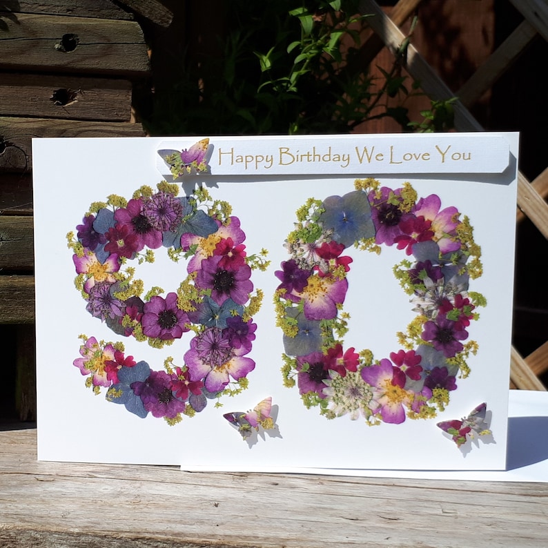 May include: A white card with the number 90 made from pressed flowers. The flowers are in shades of purple, pink, and blue. The card has the text "Happy Birthday We Love You" printed on it.