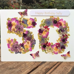 May include: A handmade birthday card with the number 50 formed from dried flowers in shades of purple, pink, and yellow. The card includes the text "Happy Birthday Sally" and decorative butterfly accents.